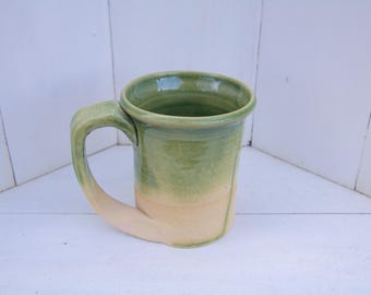 May include: A green and cream ceramic mug with a rounded handle. The mug has a glaze that creates a gradient effect from green to cream.