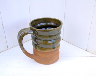 May include: A handmade ceramic mug with a green and brown glaze. The mug has a wide, rounded base and a slightly flared rim. The handle is attached to the side of the mug.