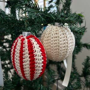以下が含まれることがあります： 緑のクリスマスツリーに吊るされた2つの手作りのクリスマスオーナメント。1つのオーナメントは赤と白のストライプで、もう1つは無地のクリーム色です。どちらのオーナメントにも、銀色の金属製のキャップと白いリボンが付いています。
