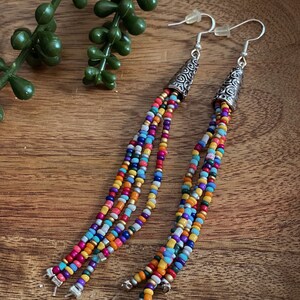 Puede incluir: Pendientes de cuentas de colores con puntas cónicas plateadas y ganchos para las orejas. Los pendientes presentan múltiples hebras de pequeñas cuentas en varios colores, incluyendo rojo, naranja, amarillo, azul y morado. Los pendientes se muestran sobre una superficie de madera.
