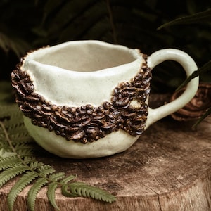 May include: A white ceramic mug with a brown textured design around the rim and handle. The mug is sitting on a wooden surface with green foliage in the background.