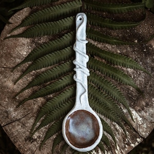 May include: A white ceramic spoon with a swirling design, resembling a vine, on the handle. The spoon has a brown glazed bowl and is resting on a wooden surface with green ferns.
