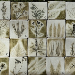 May include: A collection of twenty square ceramic tiles arranged in a grid, each featuring a unique botanical imprint in dark olive green on a cream background. Each tile is labelled with a letter from A to T, showcasing various leaves, grasses, and wildflowers.
