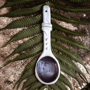 May include: A white ceramic spoon with a dark brown glaze on the inside. The spoon has a carved design on the handle and is resting on a wooden surface with green ferns.