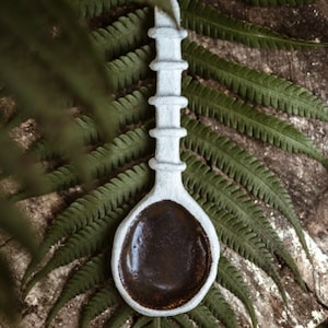 May include: A white ceramic spoon with a brown glaze inside. The spoon has a long handle with raised rings. The spoon is resting on a wooden surface with green ferns.