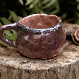 Puede incluir: Una taza de cerámica marrón oscuro con una superficie rugosa y texturizada. La taza tiene una asa ancha y redondeada y un borde ligeramente acampanado.