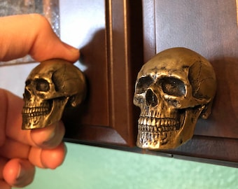 May include: Two metallic, brass-toned cabinet knobs shaped like realistic human skulls. Each knob features detailed anatomical structures including eye sockets, a nasal cavity, and teeth, with a weathered, antique finish.