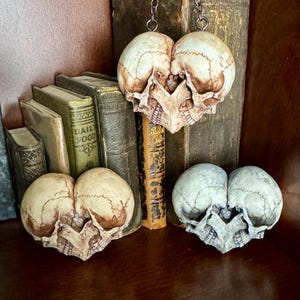 Three double skull hearts among old books, two antiqued ivory and one pale blue-gray, one hanging from a chain