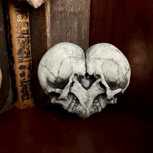 Pale gray-ivory double skull heart standing upright in front of old books, no chain