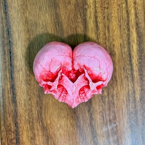 Pale pink double skull heart sculpture with red accents lying on a wooden table