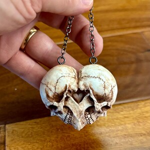 A hand holding an antiqued ivory double skull heart up by its dark chain over a wooden surface