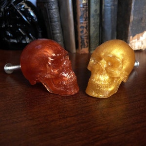 May include: Two decorative skull cabinet knobs. One is a translucent orange-red color, and the other is gold. Both have a metallic sheen and are mounted on silver screws. The knobs are likely made of resin and are designed for furniture.