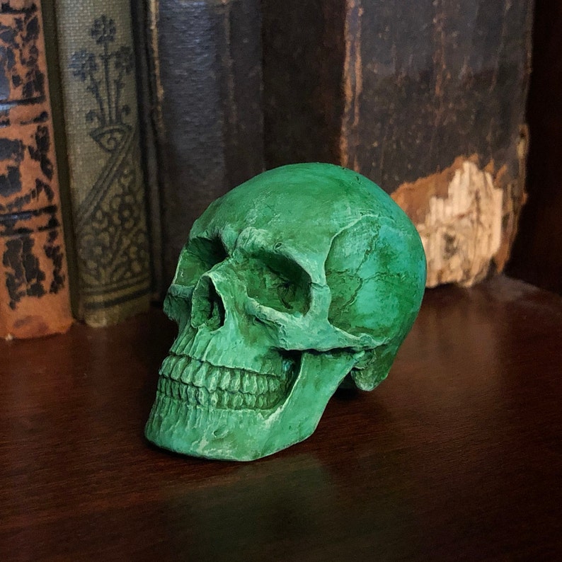 K&ouml;nnte beinhalten: Eine gr&uuml;ne dekorative Sch&auml;del-Figur mit detaillierten Gesichtsz&uuml;gen, einschlie&szlig;lich Z&auml;hne und Augenh&ouml;hlen. Der Sch&auml;del befindet sich auf einer dunklen Holzoberfl&auml;che, mit antiken B&uuml;chern im Hintergrund.