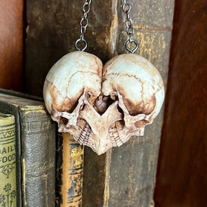 Close-up of an antiqued ivory double skull heart with brown shading hanging by its chain in front of old books