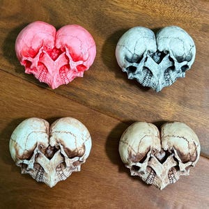 Four double skull heart sculptures on dark wood - pink, blue-gray, aged ivory, and antiqued white