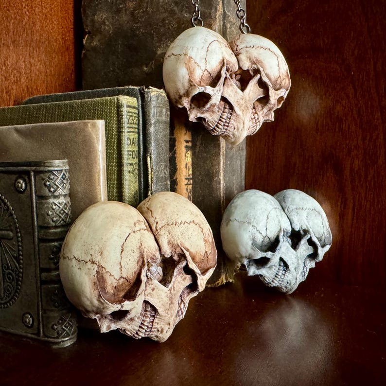 Three double skull hearts in antiqued ivory and pale blue-gray beside old books, one hanging from a chain