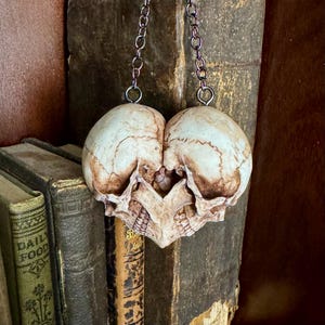 Antiqued ivory double skull heart with brown shading, hanging by a dark chain in front of old books