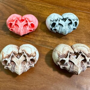 Four skull heart sculptures in pink, blue-gray, ivory, and white arranged on a wooden table