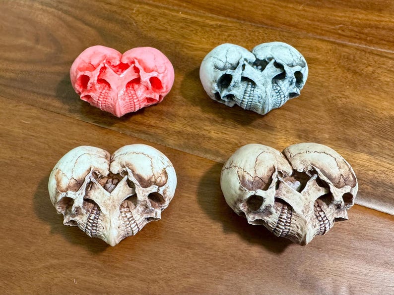 Four skull heart sculptures in pink, blue-gray, ivory, and white arranged on a wooden table
