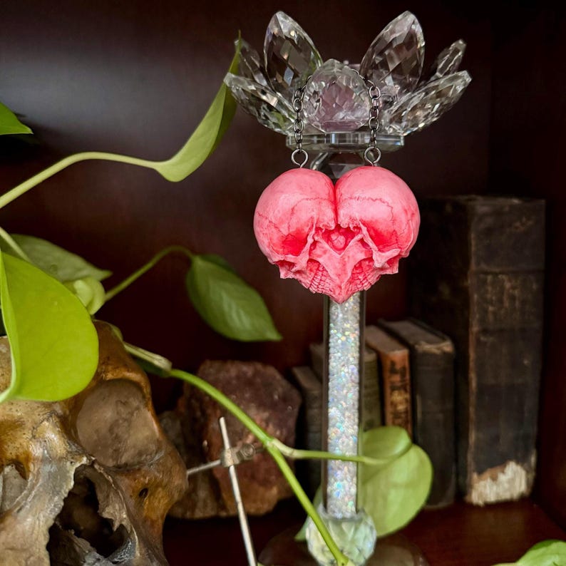 Pink skull heart sculpture standing upright below a crystal candle holder among books and vines
