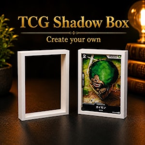 Può includere: Due cornici rettangolari bianche tipo shadow box per carte collezionabili. Una cornice è vuota, l'altra contiene una carta TCG di One Piece con il personaggio Gaimon. Il testo TCG Shadow Box Create your own è visibile in caratteri dorati.