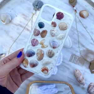 Puede incluir: Funda de teléfono blanca decorada con varias conchas marinas en tonos beige, marrón y morado. La funda está sujeta por una mano con esmalte de uñas azul. Otras conchas marinas y joyas están dispersas en una bandeja blanca.