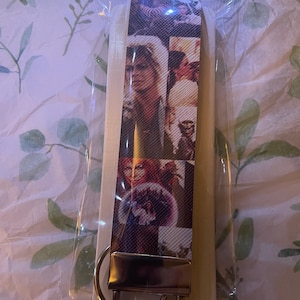May include: A rectangular fabric keychain strap featuring a collage of scenes from the movie Labyrinth, including characters like Jareth the Goblin King. The strap is attached to a silver-toned metal clasp and a circular split ring, all enclosed in a clear plastic sleeve.