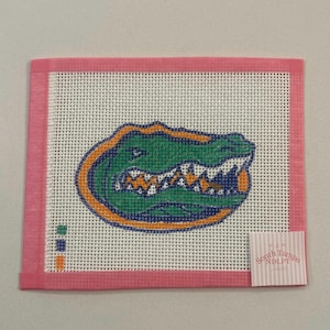 May include: A needlepoint canvas featuring a green alligator head with orange and blue accents. The alligator is centered on a white mesh background, framed by a pink border. A small tag with text is in the lower right corner.
