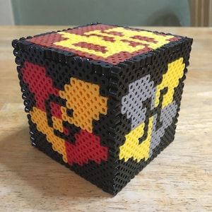 May include: A cube-shaped bead craft with a black border and a design of the Hogwarts house crests. The crests are in red, yellow, and gray.