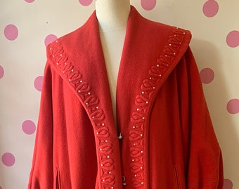 Puede incluir: Un abrigo largo de lana roja con un cuello ancho y un ribete decorativo de cuentas blancas. El abrigo tiene una silueta larga y fluida y es perfecto para una ocasión especial.