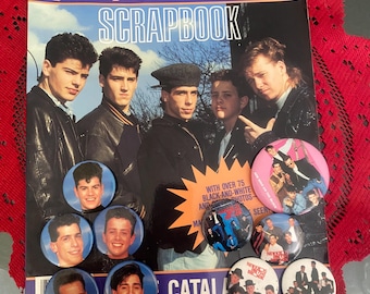 Puede incluir: Un álbum de recortes vintage de "New Kids on the Block" con una foto del grupo. Varios botones con imágenes de la banda están dispersos alrededor del álbum. El título "SCRAPBOOK" y "CATALOG" es visible.