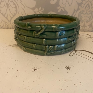 Puede incluir: Una maceta de cerámica verde con un diseño de estilo bambú. La maceta redonda presenta bandas horizontales que imitan los tallos de bambú. El interior es de color beige claro y el exterior es de color verde brillante.
