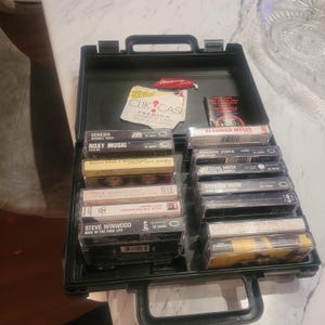 Może przedstawiać: Czarna walizka na kasety wypełniona kasetami muzycznymi w stylu vintage. Walizka jest otwarta, ukazując rzędy kaset z różnymi nazwami artystów i tytułami albumów. Widoczni są m.in. Genesis, Roxy Music i Depeche Mode. Widoczna jest wkładka papierowa.