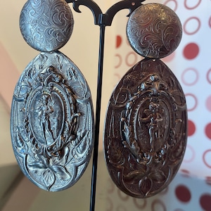 Puede incluir: Un par de pendientes llamativos plateados. Cada pendiente presenta una parte superior redonda con un patrón de remolino y un colgante ovalado con un relieve detallado de una figura dentro de un marco floral. Los pendientes se exhiben en un soporte negro.