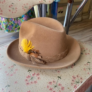 May include: A tan felt fedora hat featuring a braided band around the crown. The hat is accented with a cluster of bright yellow and brown feathers tucked into the side of the band, which also includes a small tied detail at the base.