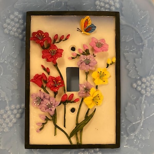 May include: A decorative light switch cover with a floral design. It features red, pink, purple, and yellow flowers, and a small butterfly. The cover has a black border and is set against a light background.