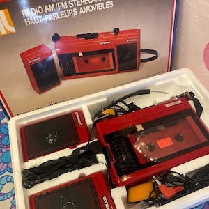 Puede incluir: Una radio cassette estéreo AM/FM roja vintage con altavoces desmontables. La radio tiene un reproductor de cassette y un dial de sintonización. La palabra "STEREO" es visible. La caja dice "RADIO AM/FM STEREO-MAGNETOCASSETTE AVEC HAUT-PARLEURS AMOVIBLES".