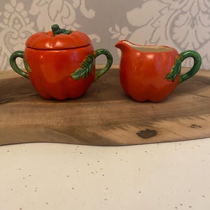 May include: A tomato-shaped ceramic sugar bowl with a lid and a creamer. The sugar bowl has a green stem and leaf handle, and the creamer has a green handle. The set is a bright red colour.