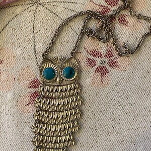 May include: A silver-toned owl pendant necklace with turquoise-colored eyes. The owl pendant has a detailed, layered feather design. The necklace chain is also silver-toned and has a textured appearance. The necklace is displayed on a floral patterned background.