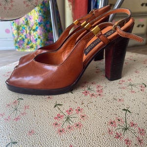 Puede incluir: Un par de zapatos de tacón destalonados de cuero color coñac con diseño peep-toe, que presentan un tacón de bloque marrón oscuro, detalles de metal dorado en las correas y un cierre de hebilla ajustable. La plantilla interior lleva el texto UNIQUE.