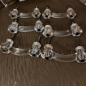 May include: A collection of clear glass cabinet pulls with faceted crystal knobs and curved handles. Each pull has a black screw for mounting. The pulls are arranged on a dark brown surface.