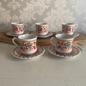 May include: Six white teacups and saucers with colourful floral designs. Each cup and saucer has a scalloped edge and a vibrant pattern of red, yellow, blue, and green flowers. The cups have small handles.