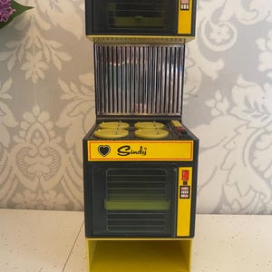 以下が含まれることがあります： 黄色と黒のヴィンテージおもちゃのキッチンセット。セットには、電子レンジ、グリル、オーブン、4つのバーナー付きコンロが含まれています。「Cindy」の文字がオーブンの前面に印刷されています。セットはプラスチック製です。
