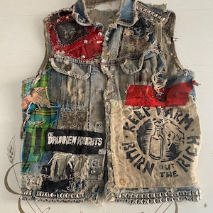 Puede incluir: Chaleco vaquero desgastado con parches, tachuelas y texto. Los parches incluyen tela roja, cuadros escoceses y un parche negro que dice "THE DRUNKEN KNIGHTS". El chaleco también presenta un parche con un gráfico de botella y las palabras "KEEP WARM BURN THE RICH OUT THE FIRE."