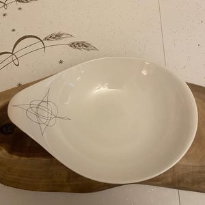 Puede incluir: Un cuenco blanco de forma ovalada con un asa en un lado. El cuenco presenta un diseño abstracto gris cerca del asa. El cuenco está colocado sobre una tabla de servir de madera.