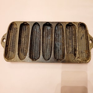 May include: A vintage cast iron cornbread pan with six individual moulds shaped like corn cobs. The pan is dark grey and has a rustic, well-used appearance.