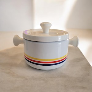 May include: White ceramic lidded pot with two side handles and a top handle. The pot is decorated with horizontal stripes in red, yellow, and blue. A kitchenware item.