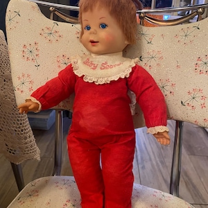 Puede incluir: Una muñeca vintage con cabello rojo recogido en un moño, vestida con un atuendo rojo y un babero que dice "Baby Secrets". La muñeca tiene ojos azules y está sentada en una silla con estampado floral. El atuendo incluye una parte superior de manga larga y pantalones.