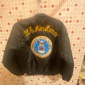 May include: Black bomber jacket with a gold embroidered "U.S. Air Force" patch and a blue and gold seal with the words "Department of the Air Force" and "United States of America".