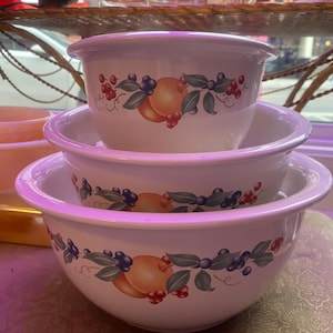 May include: A trio of white ceramic mixing bowls, each adorned with a fruit design featuring peaches, blueberries, and red berries. The bowls are stacked, displaying their different sizes and rounded rims. Measurements are in centimeters.